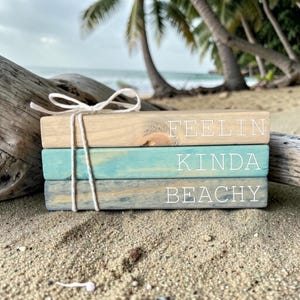 May include: Stacked wooden blocks with the words "FEELIN KINDA BEACHY" painted on them. The top block is natural wood, the middle is turquoise, and the bottom is gray-blue. A white string is tied around the blocks, and they rest on sand.