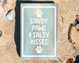 May include: A light blue wooden sign with white text that reads "Sandy Paws & Salty Kisses". The sign has two white paw prints on it.