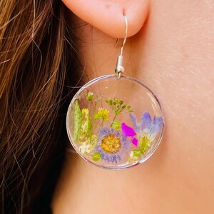 Puede incluir: Pendientes circulares transparentes con flores secas en tonos morados, amarillos y verdes. Los pendientes cuelgan de ganchos plateados. Se ve un collar de gargantilla de encaje negro.