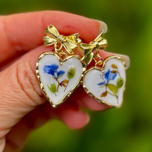 May include: Two gold heart-shaped earrings with a white background and blue and green pressed flowers inside. The earrings have a gold bow at the top.