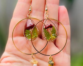 Op de afbeelding: Goudkleurige oorringen. Elke oorbel heeft een druppelvormige hars hanger met een rood en groen botanisch ontwerp. Eén oorbel heeft een kleine citrien- en parelbedel, de andere heeft een gouden eikelbedel.