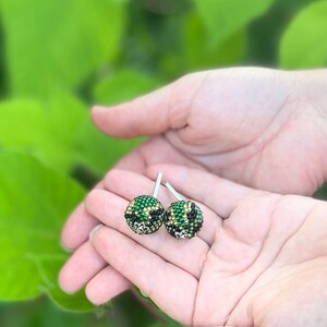 Może przedstawiać: Para okrągłych kolczyków z zielonych i złotych koralików. Kolczyki są trzymane w dłoni.