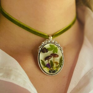 K&ouml;nnte beinhalten: Eine gr&uuml;ne Samt-Chokerkette mit einem silbernen ovalen Anh&auml;nger, der ein gepresstes Pilz- und Blumendesign zeigt.