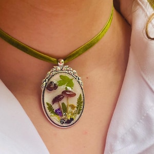 K&ouml;nnte beinhalten: Eine gr&uuml;ne Samt-Chokerkette mit einem silbernen ovalen Anh&auml;nger mit einem gepressten Blumen- und Pilzdesign.