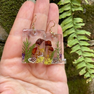 May include: A pair of gold dangle earrings with clear resin squares containing pressed flowers, mushrooms, and seashells.