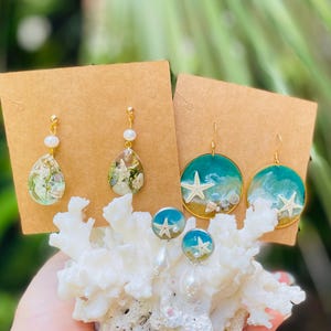 Op de afbeelding: Een collectie handgemaakte oorbellen met oceaan-thema ontwerpen. De oorbellen bevatten druppelvormige harsstukken met ingebedde schelpen en parels, en ronde oorbellen met zeesterren en oceaangolfontwerpen. De oorbellen worden gepresenteerd op bruine kaarten.
