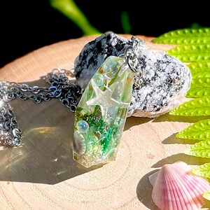 K&ouml;nnte beinhalten: Ein klarer Harzanh&auml;nger mit einem Seestern und gr&uuml;nen Einschl&uuml;ssen, an einer Silberkette. Der Anh&auml;nger liegt auf einem kleinen Stein, mit Muscheln und einem Farnwedel in der N&auml;he. Der Anh&auml;nger ist etwa 5 cm lang.