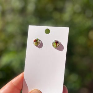 May include: A pair of small, round earrings with a clear resin base and a pressed flower design. The flowers are green, brown, and white.