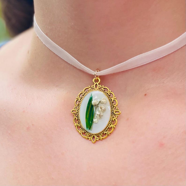 Pode incluir: Um colar com um pingente oval dourado em uma fita branca. O pingente apresenta um fundo branco com uma folha verde e flores brancas de l&iacute;rio do vale.