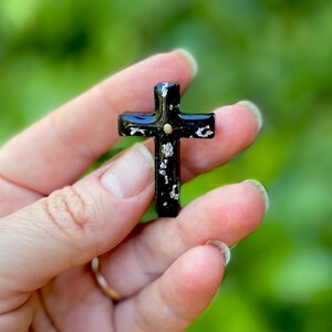 May include: A small black cross with silver glitter embedded in the resin. The cross is held in a hand.