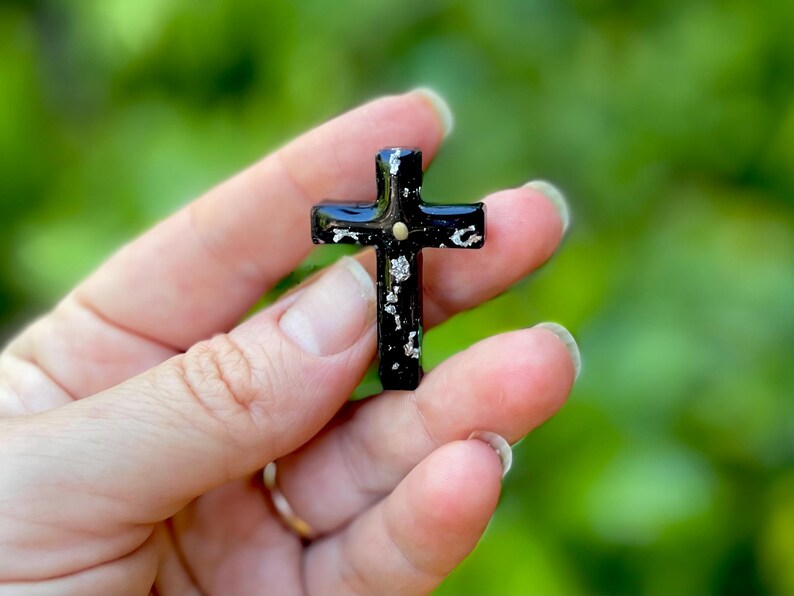 May include: A small black cross with silver glitter embedded in the resin. The cross is held in a hand.
