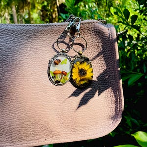 May include: Two keyrings with botanical designs hang from a pink purse. One keyring features mushrooms and greenery, the other a yellow sunflower. The keyrings have silver-coloured metal frames and key rings.