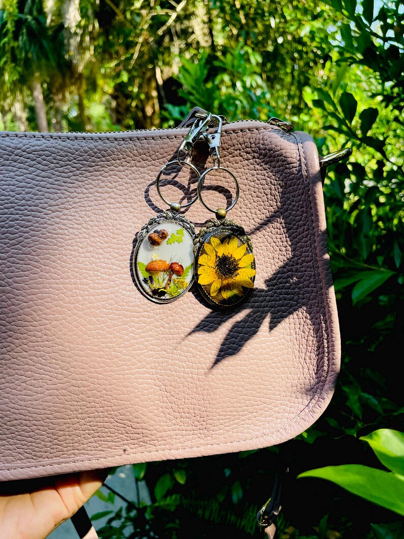May include: Two keyrings with botanical designs hang from a pink purse. One keyring features mushrooms and greenery, the other a yellow sunflower. The keyrings have silver-coloured metal frames and key rings.