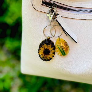 May include: Two keyrings with floral designs. One features a sunflower in a bronze-coloured frame, the other a yellow teardrop shape with a flower and green accents. They hang from a silver clasp attached to a white handbag.