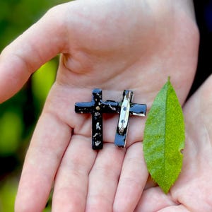 May include: Two black cross pins with a silver pin back, held in the palm of a hand. A green leaf is also in the hand.