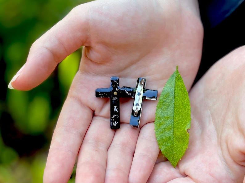 May include: Two black cross pins with a silver pin back, held in the palm of a hand. A green leaf is also in the hand.