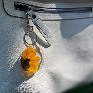 May include: A sunflower keyring encased in clear resin, attached to a silver-tone metal ring and clasp. The keyring is set against a white leather bag with a zip. The sunflower has yellow petals and a dark brown centre.
