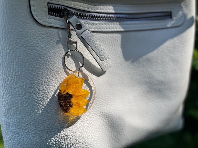 May include: A sunflower keyring encased in clear resin, attached to a silver-tone metal ring and clasp. The keyring is set against a white leather bag with a zip. The sunflower has yellow petals and a dark brown centre.