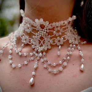 May include: A white beaded choker necklace with a delicate, intricate design. The necklace features multiple strands of beads and a central piece with a floral motif. The necklace is accented with small pearl-like beads.