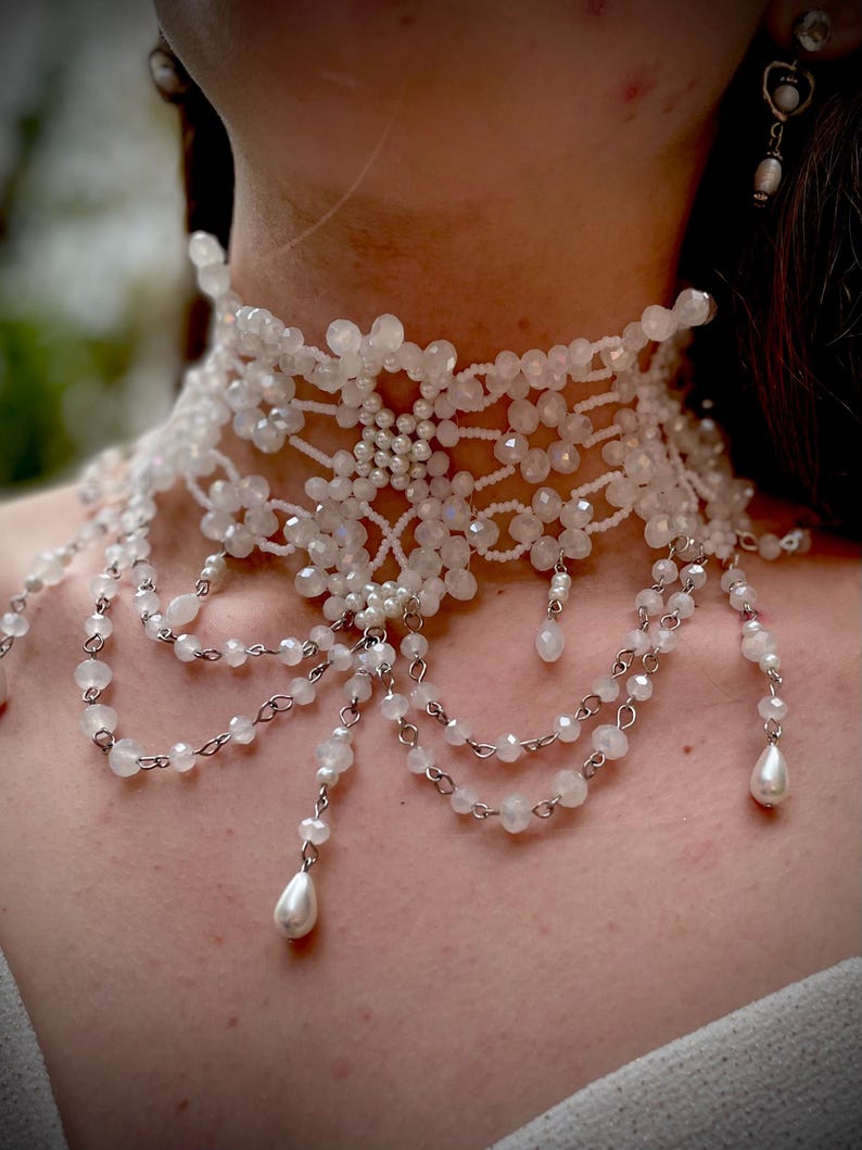 May include: A white beaded choker necklace with a delicate, intricate design. The necklace features multiple strands of beads and a central piece with a floral motif. The necklace is accented with small pearl-like beads.