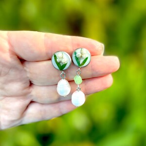 May include: Silver-toned dangle earrings featuring round, clear resin pendants with lily of the valley flowers. Each earring has a white pearl and green bead drop. The earrings are held in a hand against a blurred green background.