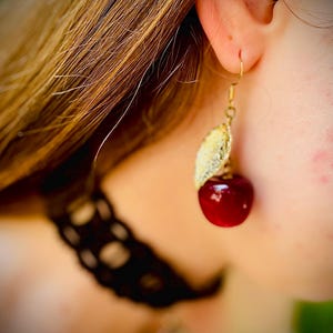 Peut inclure: Boucle d'oreille dor&eacute;e avec un motif de feuille et une cerise rouge brillante. La boucle d'oreille est port&eacute;e &agrave; l'oreille d'une personne aux cheveux ch&acirc;tain clair. Un bijou unique en forme de cerise.
