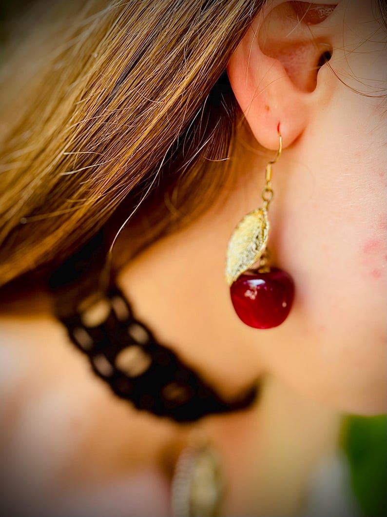 Peut inclure: Boucle d'oreille dor&eacute;e avec un motif de feuille et une cerise rouge brillante. La boucle d'oreille est port&eacute;e &agrave; l'oreille d'une personne aux cheveux ch&acirc;tain clair. Un bijou unique en forme de cerise.