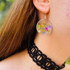 Puede incluir: Pendientes circulares transparentes con flores secas en tonos morados, amarillos y verdes. Los pendientes cuelgan de ganchos plateados. Debajo de los pendientes, se ve un collar de gargantilla de encaje negro.