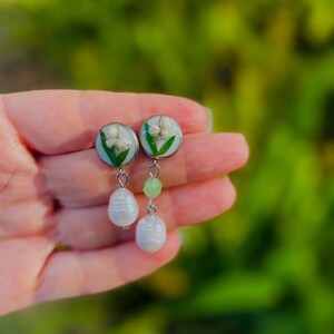 May include: A pair of earrings featuring white flowers encased in clear resin, with a silver-colored metal backing. Each earring has a white, teardrop-shaped pearl and a green bead dangling below. The background is a blurred green.