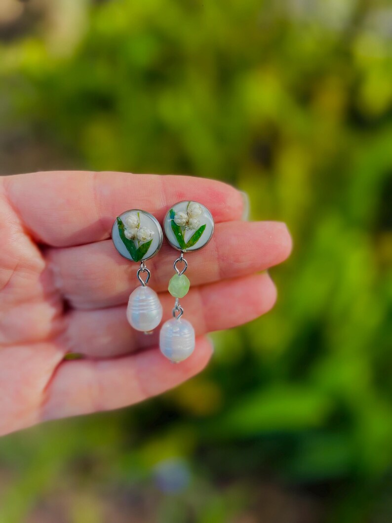 May include: A pair of earrings featuring white flowers encased in clear resin, with a silver-colored metal backing. Each earring has a white, teardrop-shaped pearl and a green bead dangling below. The background is a blurred green.