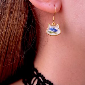 Puede incluir: Pendiente dorado con forma de gato con fondo blanco y dise&ntilde;o floral azul. El pendiente se lleva en la oreja de una persona. El pendiente tiene cierre de gancho.