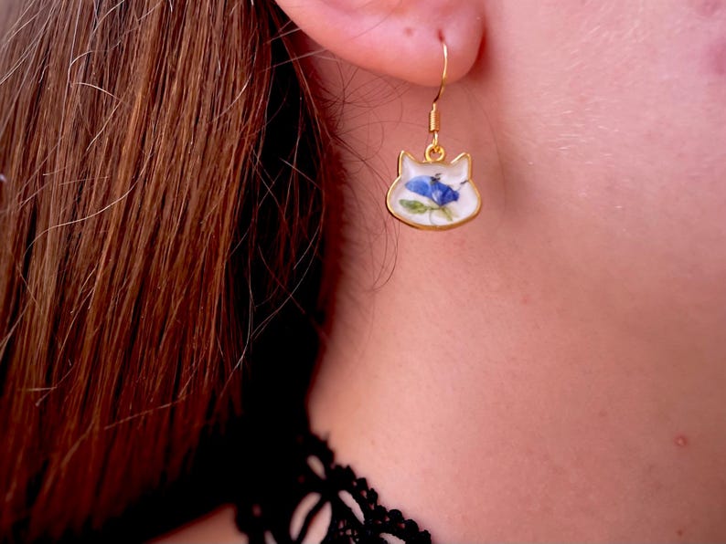 Puede incluir: Pendiente dorado con forma de gato con fondo blanco y dise&ntilde;o floral azul. El pendiente se lleva en la oreja de una persona. El pendiente tiene cierre de gancho.