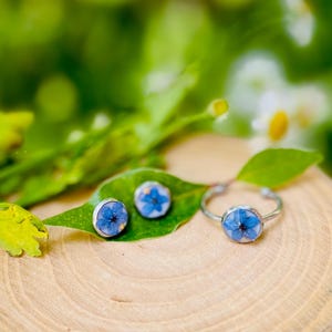 K&ouml;nnte beinhalten: Ein Set aus silbernem Schmuck mit einer blauen Blume, die in jedes St&uuml;ck gepresst ist. Das Set enth&auml;lt einen Ring und zwei Ohrringe.