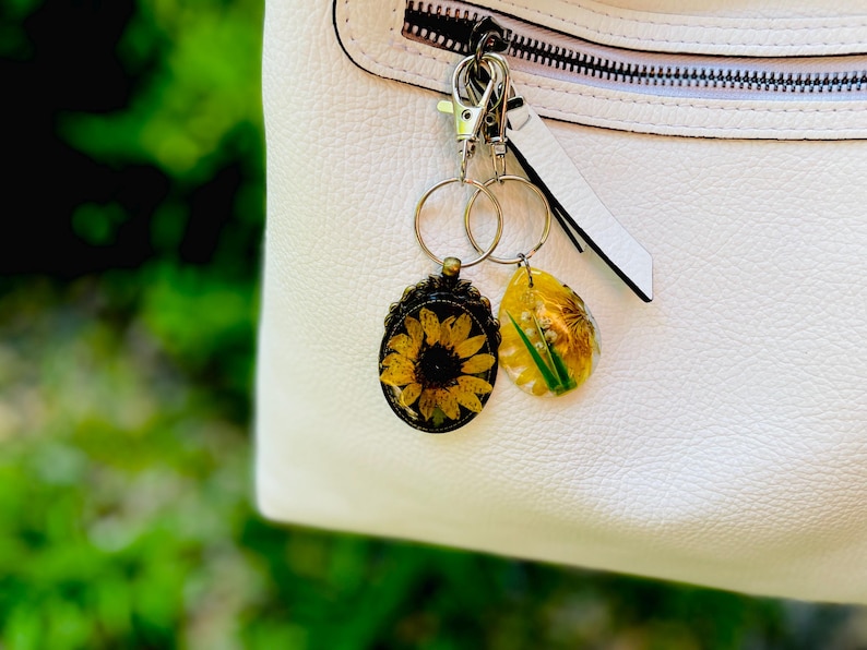 May include: Two keyrings with floral designs. One features a sunflower in a bronze-coloured frame, the other a yellow teardrop shape with a flower and green accents. They hang from a silver clasp attached to a white handbag.