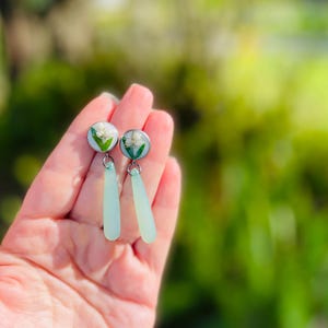 May include: A pair of earrings featuring small, round, silver-toned studs with white flowers encased in clear resin. Each earring has a light blue, teardrop-shaped dangle. The earrings are held in a hand against a blurred green background.