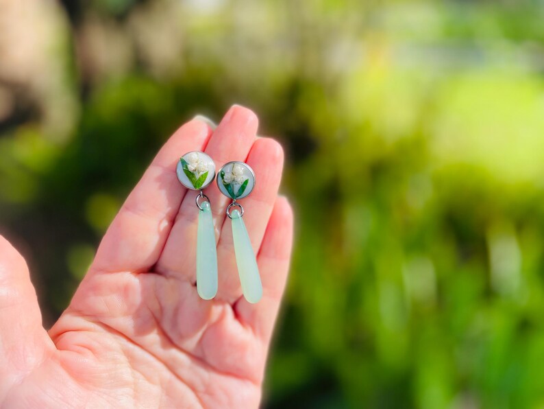May include: A pair of earrings featuring small, round, silver-toned studs with white flowers encased in clear resin. Each earring has a light blue, teardrop-shaped dangle. The earrings are held in a hand against a blurred green background.