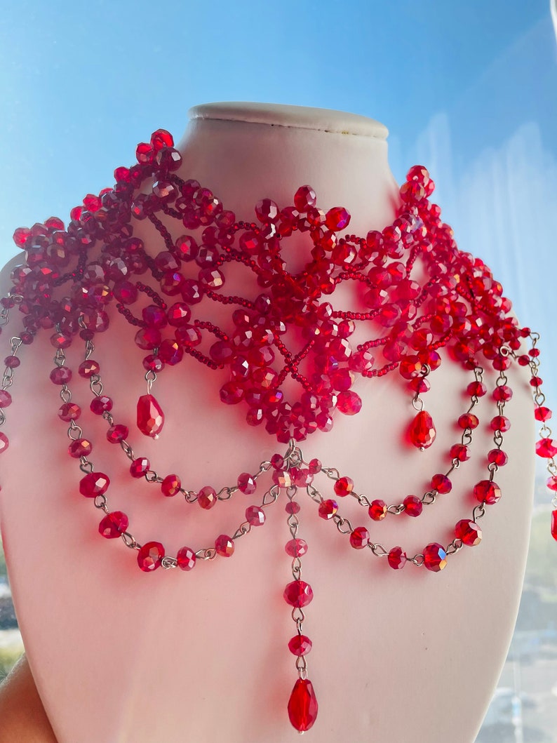 May include: A red beaded choker necklace with a delicate, intricate design. The necklace features multiple strands of beads and dangling drops, creating a layered and elegant look.