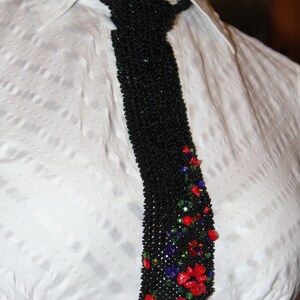 May include: A black beaded tie with a floral design. The tie is adorned with red, green, and blue beads, creating a colorful accent against the black background. The tie is worn over a white collared shirt.