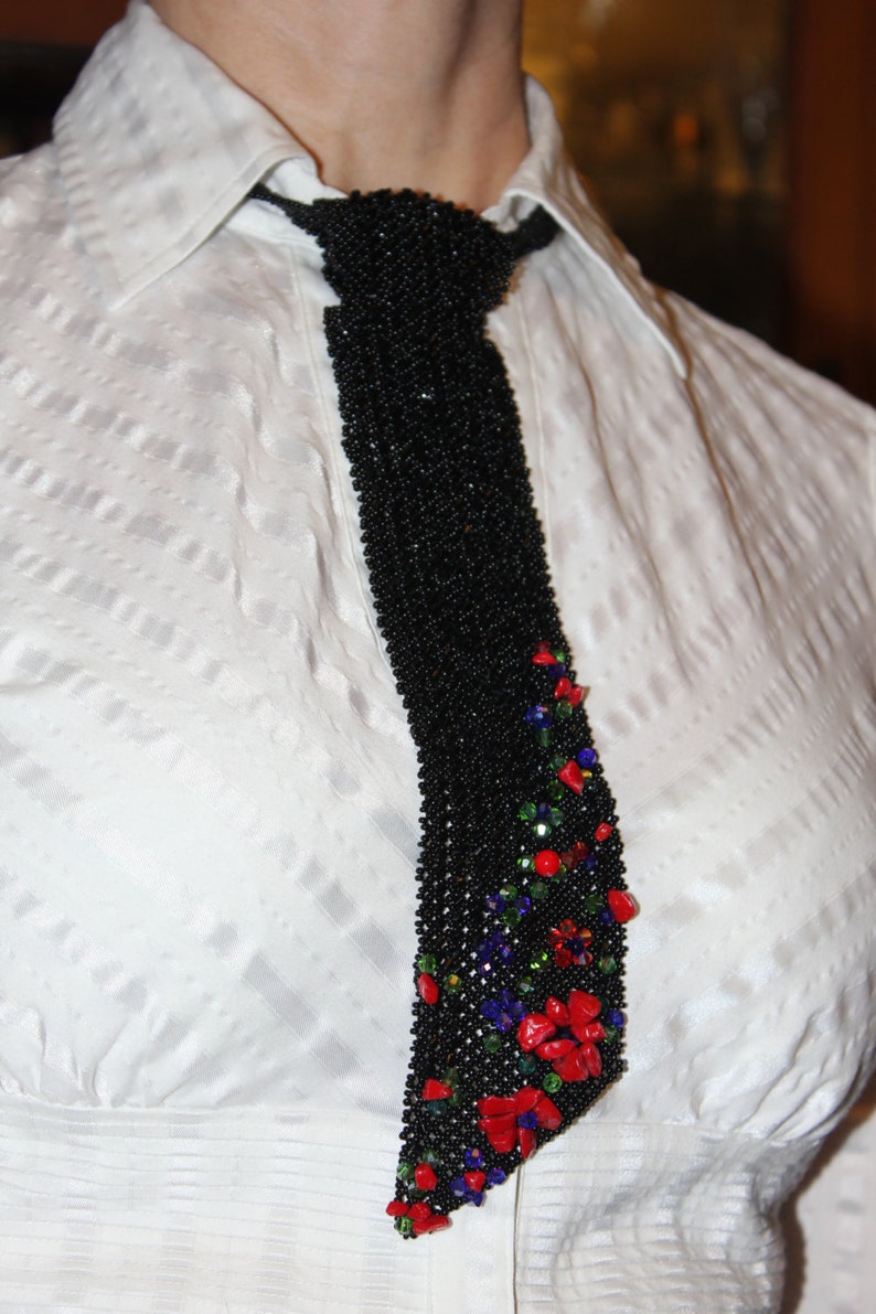 May include: A black beaded tie with a floral design. The tie is adorned with red, green, and blue beads, creating a colorful accent against the black background. The tie is worn over a white collared shirt.