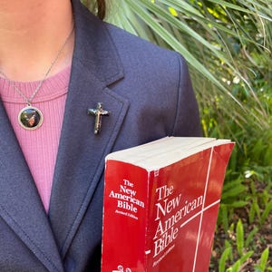 May include: A person wearing a dark blue blazer and a pink shirt with a silver chain necklace and a small round pendant. The person is holding a red book titled "The New American Bible, Revised Edition".
