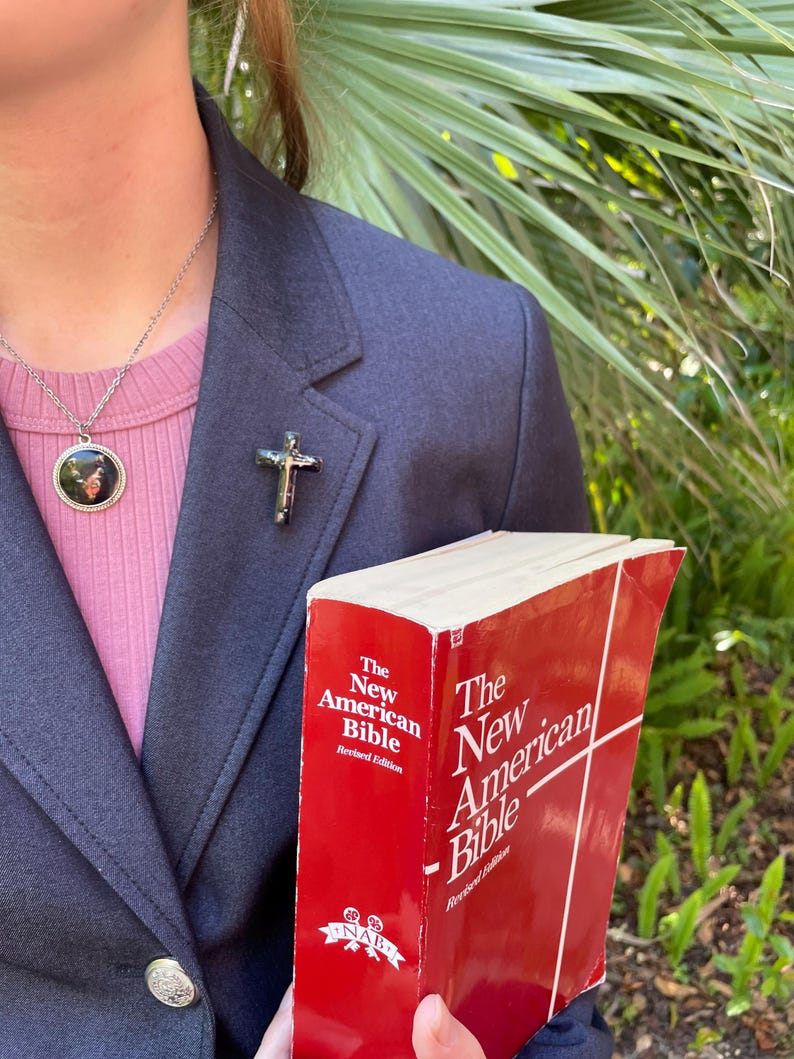 May include: A person wearing a dark blue blazer and a pink shirt with a silver chain necklace and a small round pendant. The person is holding a red book titled "The New American Bible, Revised Edition".