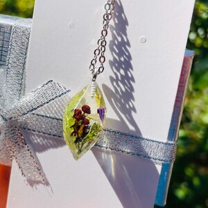 May include: A silver chain necklace with a clear resin pendant featuring pressed flowers and leaves. The pendant is shaped like a teardrop and has a light green hue.