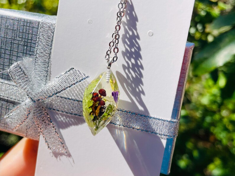May include: A silver chain necklace with a clear resin pendant featuring pressed flowers and leaves. The pendant is shaped like a teardrop and has a light green hue.