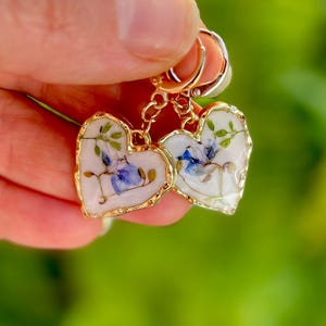 May include: A pair of gold heart-shaped earrings with pressed flowers inside. The flowers are blue, green, and white.