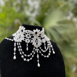 May include: A white beaded choker necklace with a delicate floral design and dangling pearl accents.