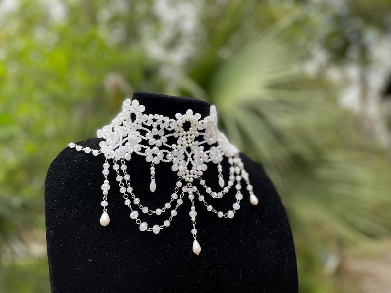 May include: A white beaded choker necklace with a delicate floral design and dangling pearl accents.