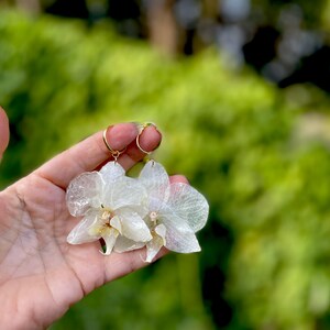 Pode incluir: Um par de brincos com p&eacute;talas de orqu&iacute;dea transl&uacute;cidas em tom branco-sujo, presas a argolas finas de metal dourado. As p&eacute;talas possuem uma textura delicada e nervurada com um centro amarelo p&aacute;lido, criando uma est&eacute;tica bot&acirc;nica leve.