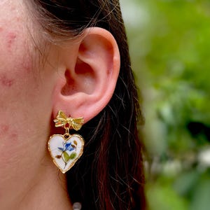 K&ouml;nnte beinhalten: Goldene herzf&ouml;rmige Ohrringe mit einem Blumenmuster und einem kleinen Schleifenanh&auml;nger. Die Ohrringe sind wei&szlig; mit blauen und gr&uuml;nen floralen Akzenten.