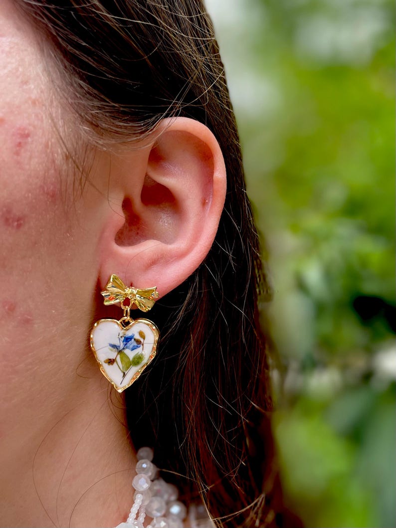 K&ouml;nnte beinhalten: Goldene herzf&ouml;rmige Ohrringe mit einem Blumenmuster und einem kleinen Schleifenanh&auml;nger. Die Ohrringe sind wei&szlig; mit blauen und gr&uuml;nen floralen Akzenten.