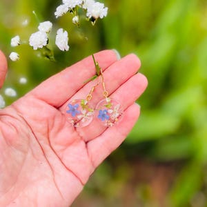 Puede incluir: Un par de pendientes de aro dorados con peque&ntilde;as flores azules y blancas prensadas en resina transparente.